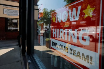 Un cartel rojo y blanco de 'Now Hiring' (Ahora Contratando) se ve a través de una ventana, con reflejos de la calle y vehículos