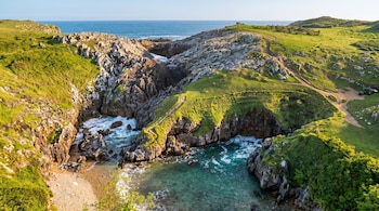 Acantilados rocosos verdes, una cala con agua turquesa y olas, una playa de arena clara, un arco natural rocoso y el mar abierto bajo cielo azul.