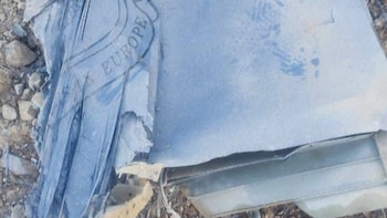 Primer plano de restos de metal de una aeronave de color gris azulado, con la palabra "EUROPE" grabada, sobre tierra y rocas pequeñas