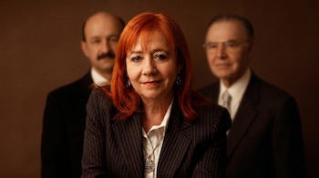 Retrato de una mujer de cabello rojizo con un traje de raya diplomática y un colgante, mirando al frente. Dos hombres borrosos aparecen detrás.