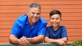 Hombre adulto y niño sonriendo, ambos con camisetas azules, apoyados sobre una barandilla verde con un fondo de pared naranja