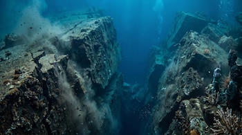 Imagen submarina de un profundo cañón rocoso con paredes escarpadas y sedimentos ascendiendo del fondo. Se observan fragmentos de roca y burbujas en el agua azul oscuro.