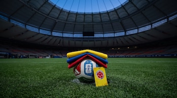 Bandera de Colombia doblada sobre balón de fútbol, gafete amarillo de la Federación Colombiana de Fútbol, césped verde y gradas vacías de estadio.