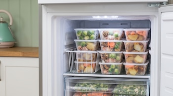 Un congelador abierto con recipientes plásticos transparentes que contienen papas, zanahorias y coles de Bruselas en sus estantes.