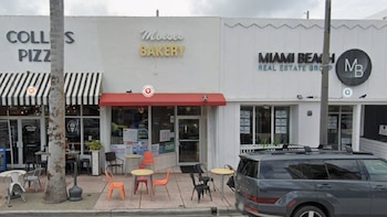 Vista frontal de una calle comercial con tres fachadas de tiendas, mesas exteriores, un poste de luz, un tronco de palmera y una SUV oscura estacionada