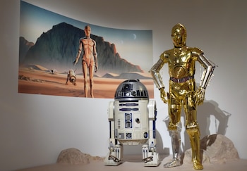 Las figuras de los droides C-3PO y R2-D2 permanecen de pie junto a una ilustración que muestra un paisaje desértico en un museo