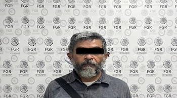 Un hombre de mediana edad, con cabello gris, barba y una barra negra sobre los ojos, posa frente a una pared decorada con los logotipos de la FGR de México.