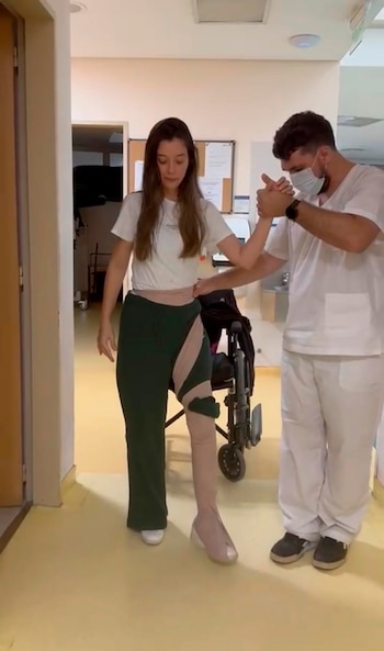 Manuela Lori, mujer con cabello largo, camina en un pasillo de hospital con un soporte en la pierna derecha, asistida por un fisioterapeuta con mascarilla