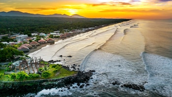 Vista aérea de una costa tropical al atardecer con olas, playa, casas con tejados de paja y montañas bajo un cielo naranja y amarillo