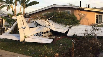 Una avioneta blanca dañada descansa sobre el techo y el garaje de una casa residencial de una planta con vegetación alrededor