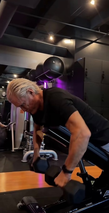 Un hombre con camisa negra y cabello rubio inclinado sobre un banco de entrenamiento sostiene dos mancuernas en un gimnasio