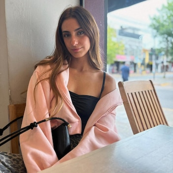 Una joven con cabello castaño y un suéter rosa se sienta en un café mirando a la cámara, con una ventana que da a una calle borrosa detrás de ella
