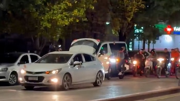 Imagen nocturna de una calle con un coche blanco de cinco puertas y maletero abierto, otro coche blanco, una furgoneta y varias motocicletas con conductores