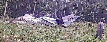 Restos de una avioneta gris y blanca con la matrícula CP-3243 esparcidos sobre hierba verde y tierra en un área boscosa, con una persona observando