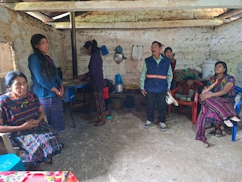 Interior de una vivienda con paredes de ladrillo blanqueado, un hombre, cuatro mujeres y dos niños. Se observan vestimentas tradicionales y objetos domésticos
