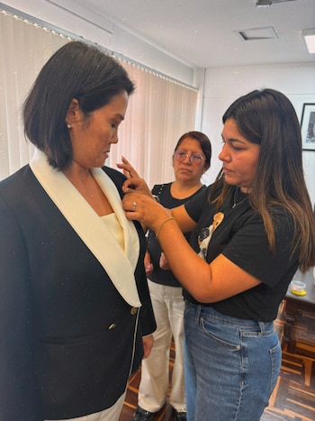 Una mujer de pelo corto y blazer negro con solapas claras es ajustada por otra mujer de pelo largo, camiseta negra y jeans, mientras una tercera observa