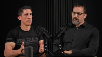 Jeff Cavaliere y Andrew Huberman sentados frente a micrófonos en un estudio oscuro con paneles verticales. Cavaliere, a la izquierda, gesticula; Huberman, a la derecha, escucha