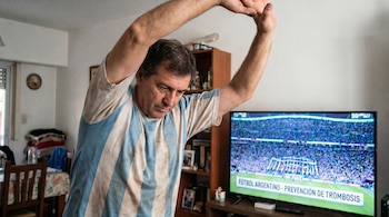Hombre de mediana edad con camiseta a rayas celestes y blancas estira los brazos sobre la cabeza mirando una televisión con un partido de fútbol en un salón.