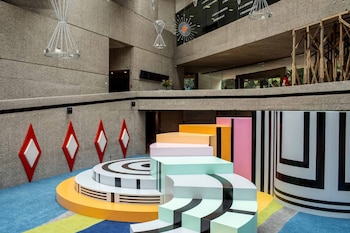 Interior de museo de concreto con alfombra azul y amarilla. Esculturas geométricas de colores pastel, podio circular, poste de rayas, rombos rojos en pared. Personas en nivel superior