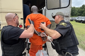 Dos oficiales de policía escoltan a un hombre esposado con uniforme naranja hacia un autobús de transporte. Otro hombre asoma su cabeza dentro del autobús