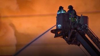 Dos bomberos en la plataforma de un camión escalera lanzan agua. El fondo es un resplandor naranja brillante de un incendio nocturno