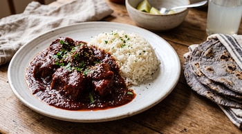 Carne en salsa roja con sésamo y arroz blanco en un plato de cerámica junto a tortillas de maíz azul y limones.