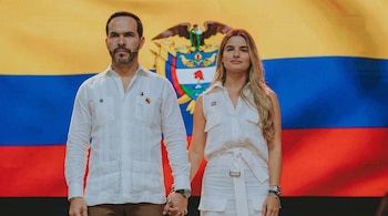 Un hombre y una mujer de pie, tomados de la mano, frente a una gran bandera de Colombia que ocupa todo el fondo. Ambos visten de blanco.