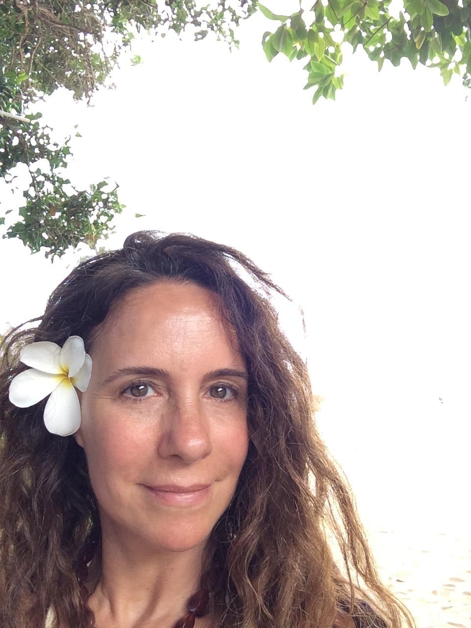 Noelle Balfour posa al aire libre con una flor blanca en el cabello bajo la sombra de unos árboles.