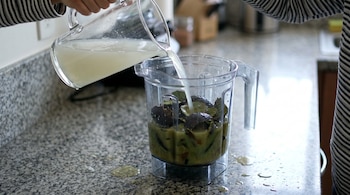Una mano vierte líquido de una jarra de vidrio a una licuadora con cáscaras de aguacate sobre una encimera de granito. Hay gotas de líquido derramadas.