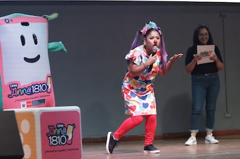 En un escenario, una persona vestida de payaso con micrófono habla junto a la mascota de la Línea ANNA 1810 y otra mujer con gafas que lee un papel