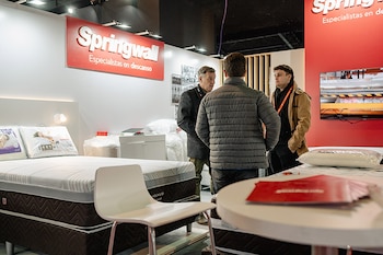 Un stand de Springwall con logotipos visibles, camas con colchones y somieres, almohadas, mesas, una silla y tres hombres hablando