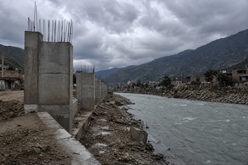 Estructuras de concreto con varillas de hierro sobresalientes en la orilla de un río, con montañas y casas al fondo bajo un cielo nublado.