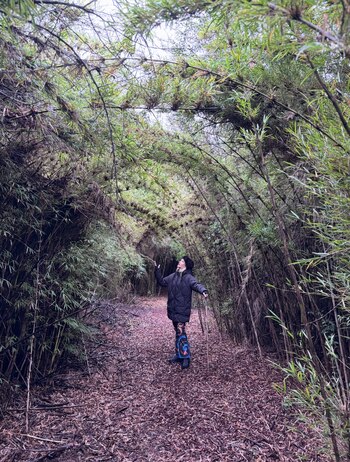 Una mujer rubia con gorro y campera negra de pie en un sendero cubierto de hojas secas, rodeado de cañas de bambú que forman un túnel, con un palo en la mano