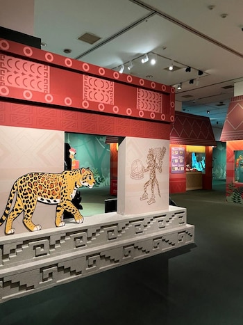 Exhibición de museo con una figura de jaguar, estructuras rojas decoradas con patrones prehispánicos y una ilustración de un guerrero antiguo
