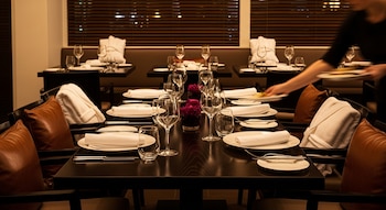 Restaurante con mesas oscuras, copas, platos, cubiertos, servilletas, cojines marrones y batas blancas en sillas. Persona borrosa asiste con el servicio.