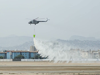 Un helicóptero militar en vuelo descarga agua desde un contenedor colgante verde sobre una pista de aterrizaje frente a edificios y cerros