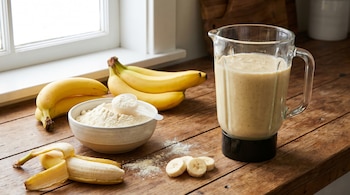 Un batido de banana en una licuadora, plátanos enteros y rebanados, y un bol con proteína en polvo sobre una mesada de madera clara, con una ventana al fondo.