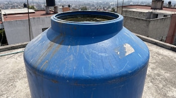Primer plano de un tinaco de plástico azul sin tapa, parcialmente lleno de agua turbia, sobre una azotea de concreto, con edificios al fondo.