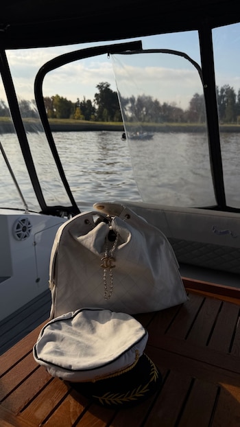 Primer plano de una cartera de lujo blanca con logo Chanel y un gorro de capitán sobre una mesa de madera en un barco. El río y árboles se ven al fondo