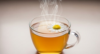 Taza de cristal transparente con líquido ámbar, una flor de manzanilla blanca y centro amarillo flotando, y vapor visible sobre un fondo neutro claro.