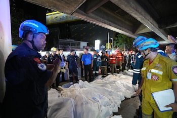 Hombres con uniformes de rescate y seguridad junto a filas de cuerpos cubiertos con sábanas blancas en una calle por la noche. Edificios y estructuras