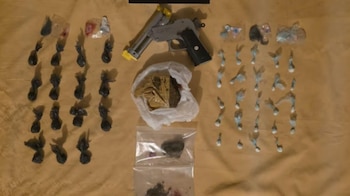 Vista aérea de varias dosis de drogas en bolsas negras y claras, un ladrillo de marihuana, y una pistola gris con detalles amarillos sobre una superficie marrón