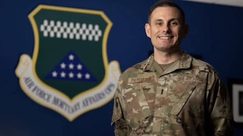 Un hombre sonriente con uniforme militar de camuflaje se encuentra delante de un escudo que lleva la inscripción Air Force Mortuary Affairs Ops