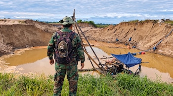 Hombre de espaldas con ropa de camuflaje y sombrero frente a una fosa de tierra con agua turbia, mangueras y una balsa con motor.
