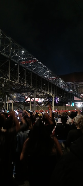 Vista nocturna de una gran multitud de personas con los brazos levantados y luces brillantes, frente a una estructura metálica de estadio