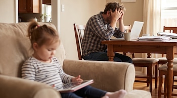 Una niña en primer plano usa una tableta en un sillón, mientras al fondo un hombre se toma la cabeza ante una computadora portátil.