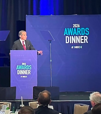 Hombre con traje y corbata roja en un podio con la inscripción 2026 AWARDS DINNER AT NECX frente a un fondo azul y asistentes