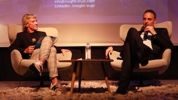 Dos hombres sentados en sillones en un escenario. Uno con traje oscuro y pantalones a cuadros, el otro con traje negro, ambos con micrófonos. Mesa y botella de agua central