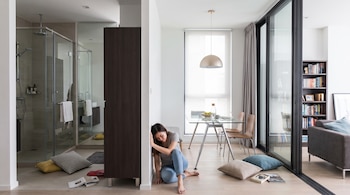 Una mujer sentada en el suelo junto a una pared en un apartamento moderno, con un baño de vidrio, una mesa, un sofá y estanterías con libros