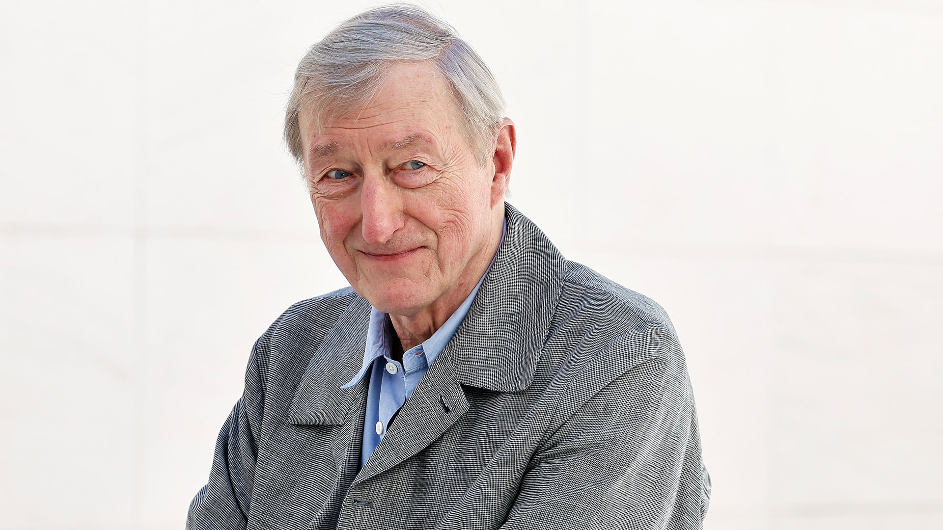 El escritor británico Julian Barnes posa para un retrato, mirando a la cámara con una chaqueta gris sobre una camisa azul clara.